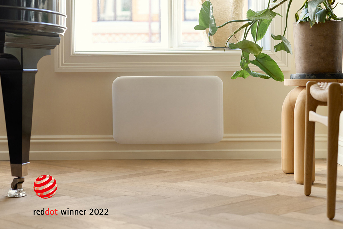 Hvit panelovn montert på vegg i et moderne rom med tregulv, stol og grønn plante. Red Dot Award-merke vises.