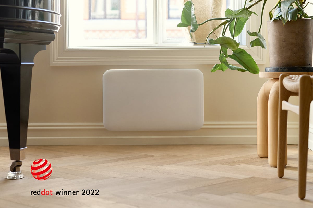 Hvit panelovn montert på vegg i et moderne rom med tregulv, stol og grønn plante. Red Dot Award-merke vises.
