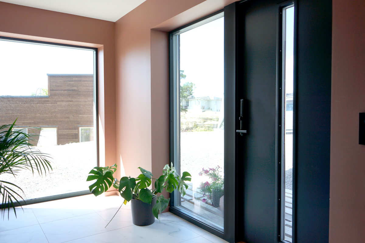 Bildet viser en moderne entré med store vindusflater som slipper inn mye dagslys. Døren er mørk og minimalistisk i design, med et smalt vindu ved siden av.