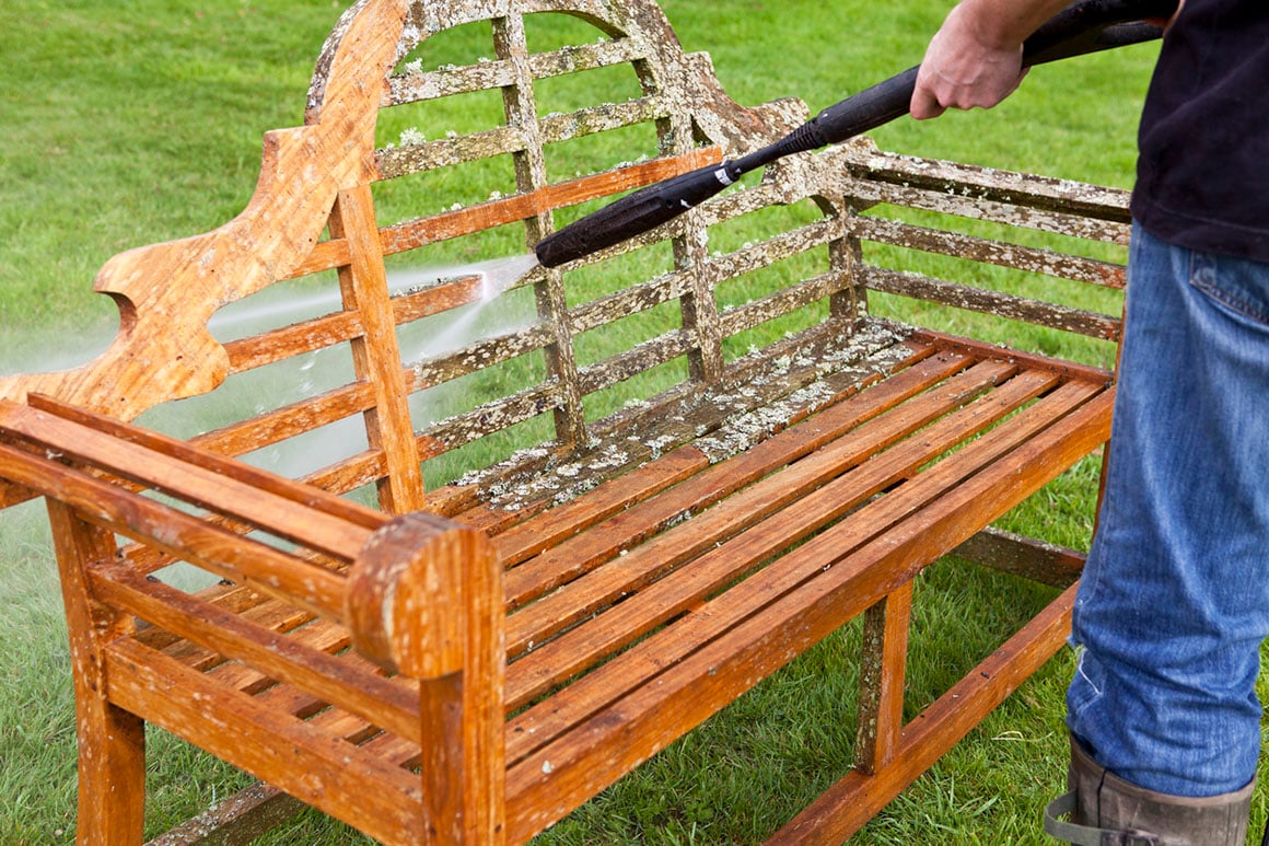Person rengjør trebenk i hagen med høytrykkspyler.