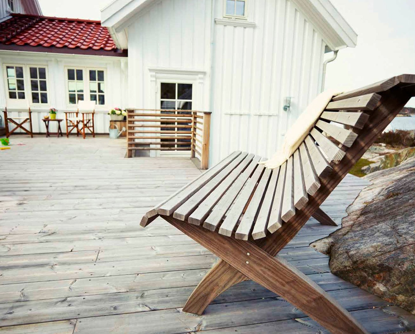 Lys treterrasse foran hvitt hus med benk og kystmiljø i bakgrunnen.