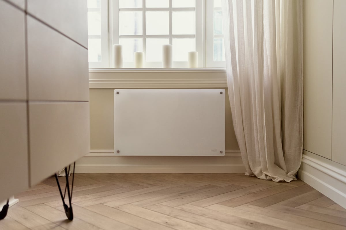 Hvit panelovn montert under vindu ved seng i lyst rom.