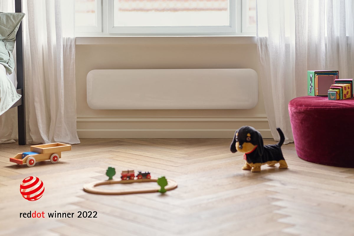 Avlang panelovn montert under vindu, med liten lekehund og andre barneleker på gulvet foran.