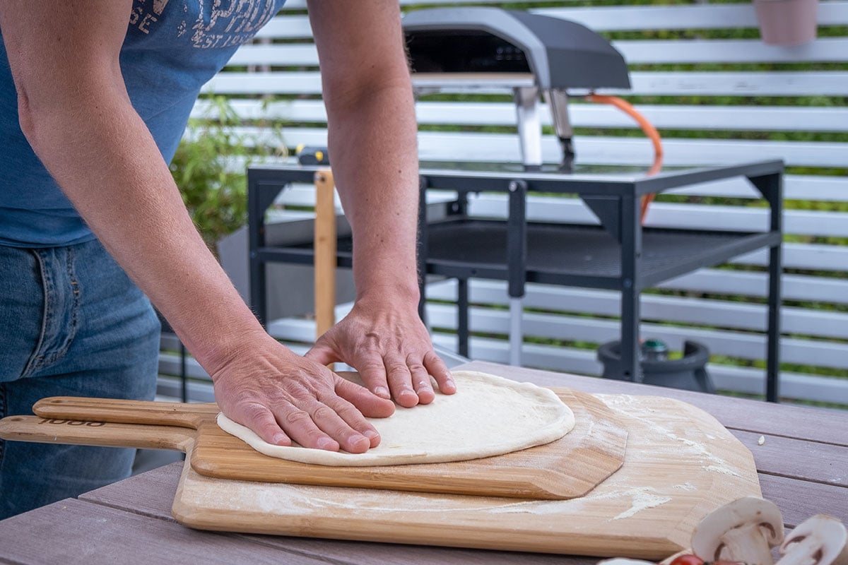 En person som trykker ut en pizzadeig på en pizzaspade. Personen står utendørs, og i bakgrunnen ser man en pizzaovn på et bord.