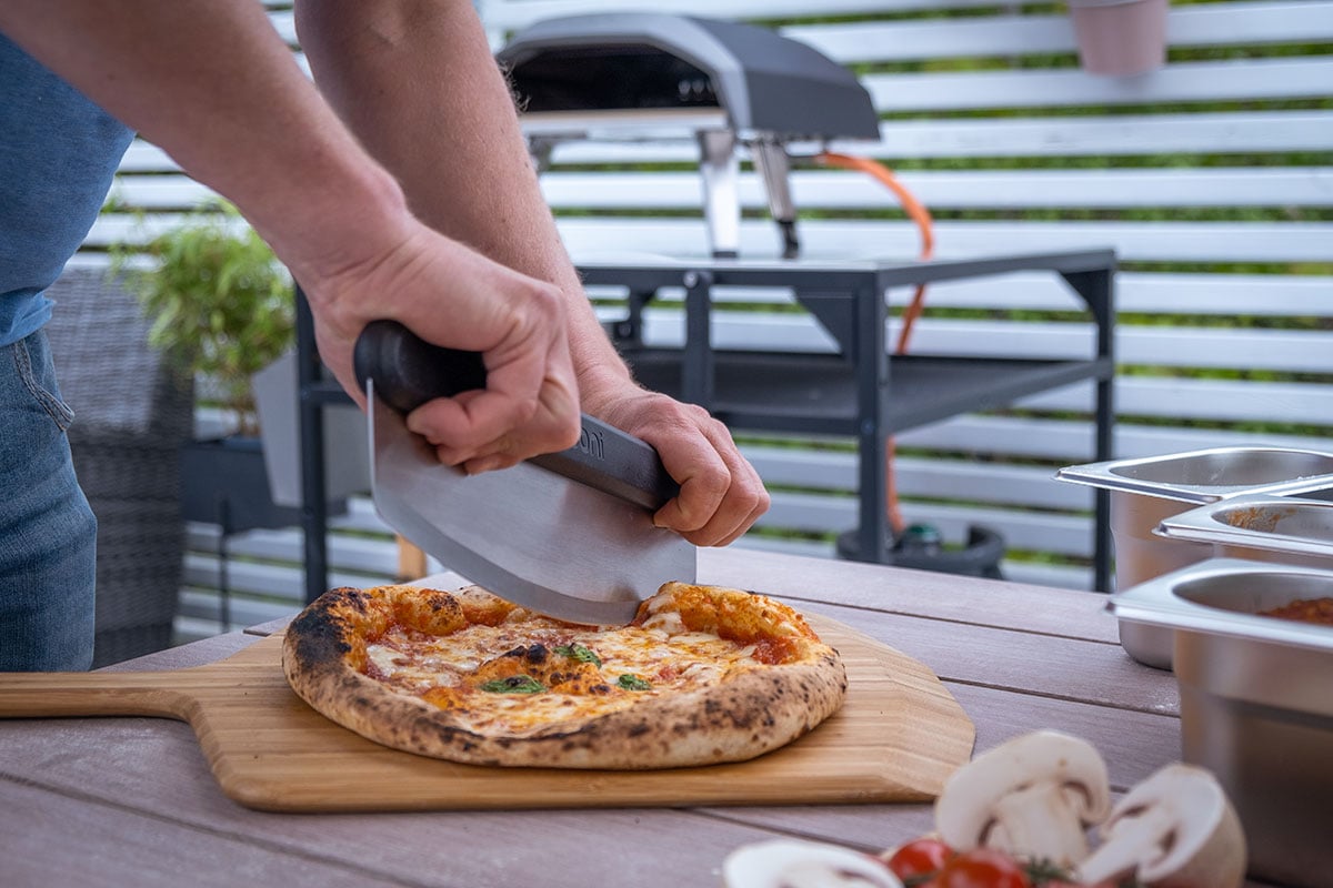 En person bruker en stor, buet kniv for å skjære opp en ferdigstekt pizza på en pizzaspade. Pizzaen ligger på et bord utendørs, og i bakgrunnen ser man en pizzaovn.