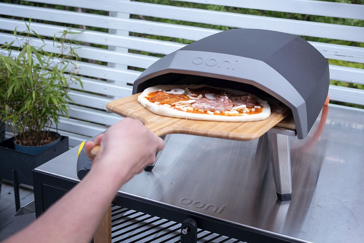 En person bruker en pizzaspade for å skyve en pizza inn i en pizzaovn. Pizzaen er allerede lagt på spaden og er klar for steking.