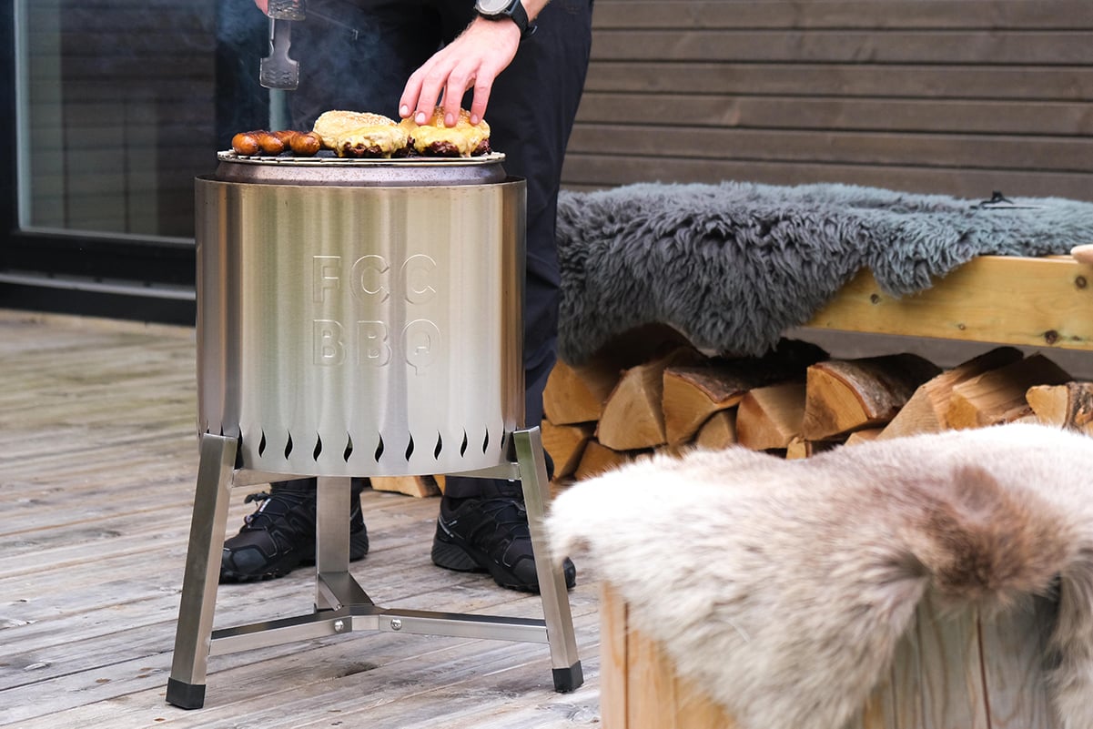 En person griller burgere og pølser på en røykfri FCC BBQ-grill på en treterrasse med ved og saueskinn.