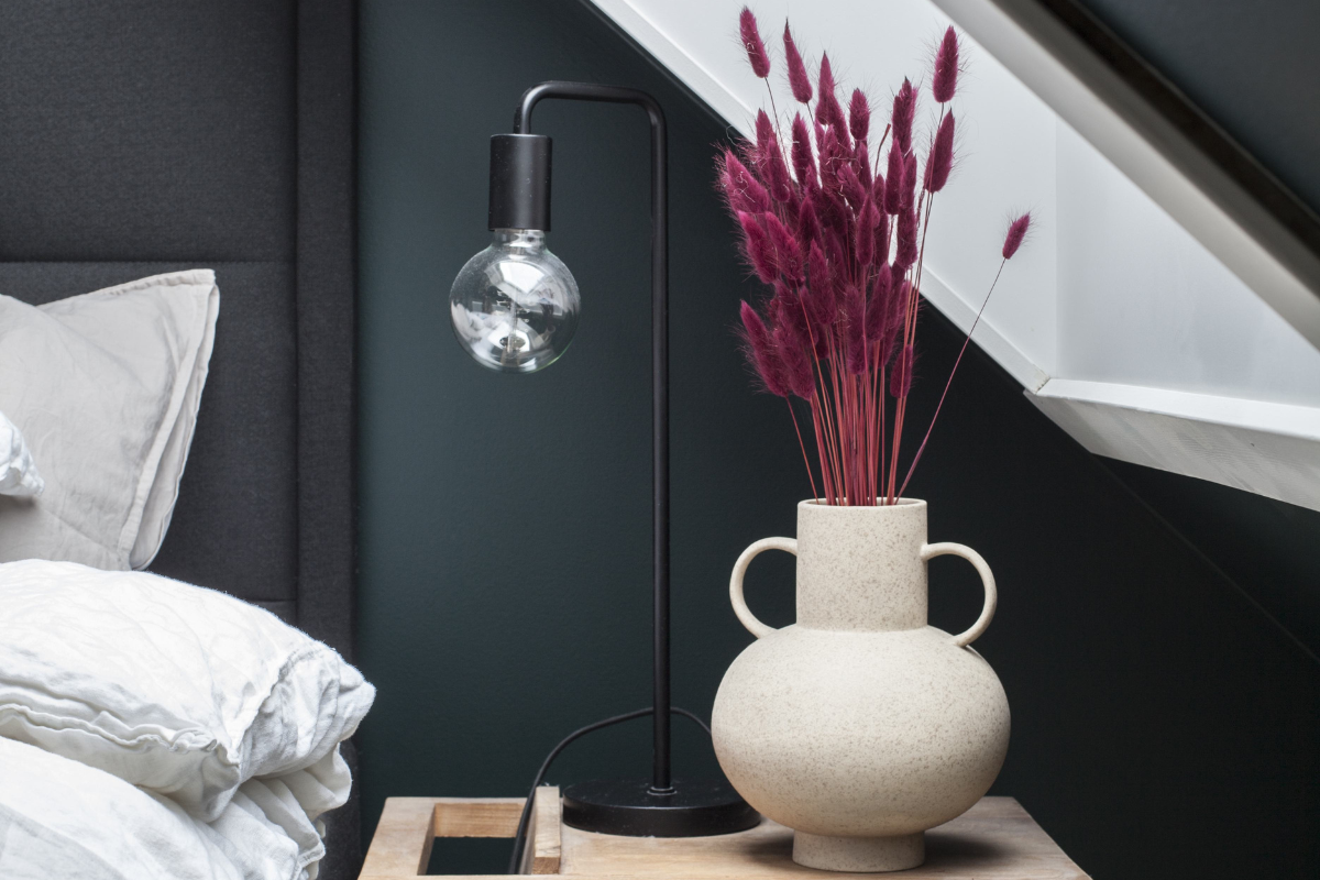 Et moderne soverom med mørke vegger i sort eller antrasitt, pyntet med vase og tørkede blomster på nattbordet.