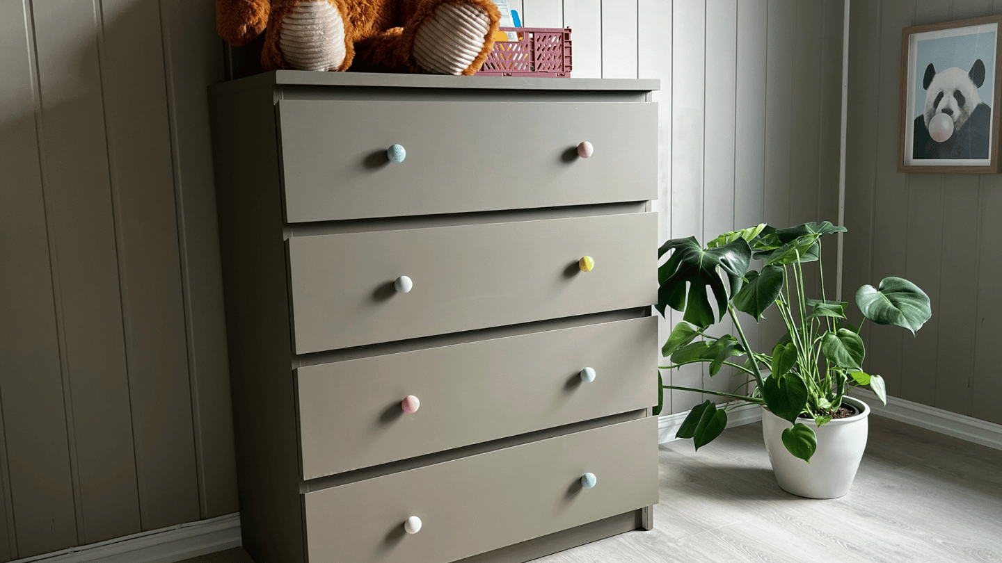 Ferdig kommode i brungrå farge med fargerike knotter og bamse på toppen.