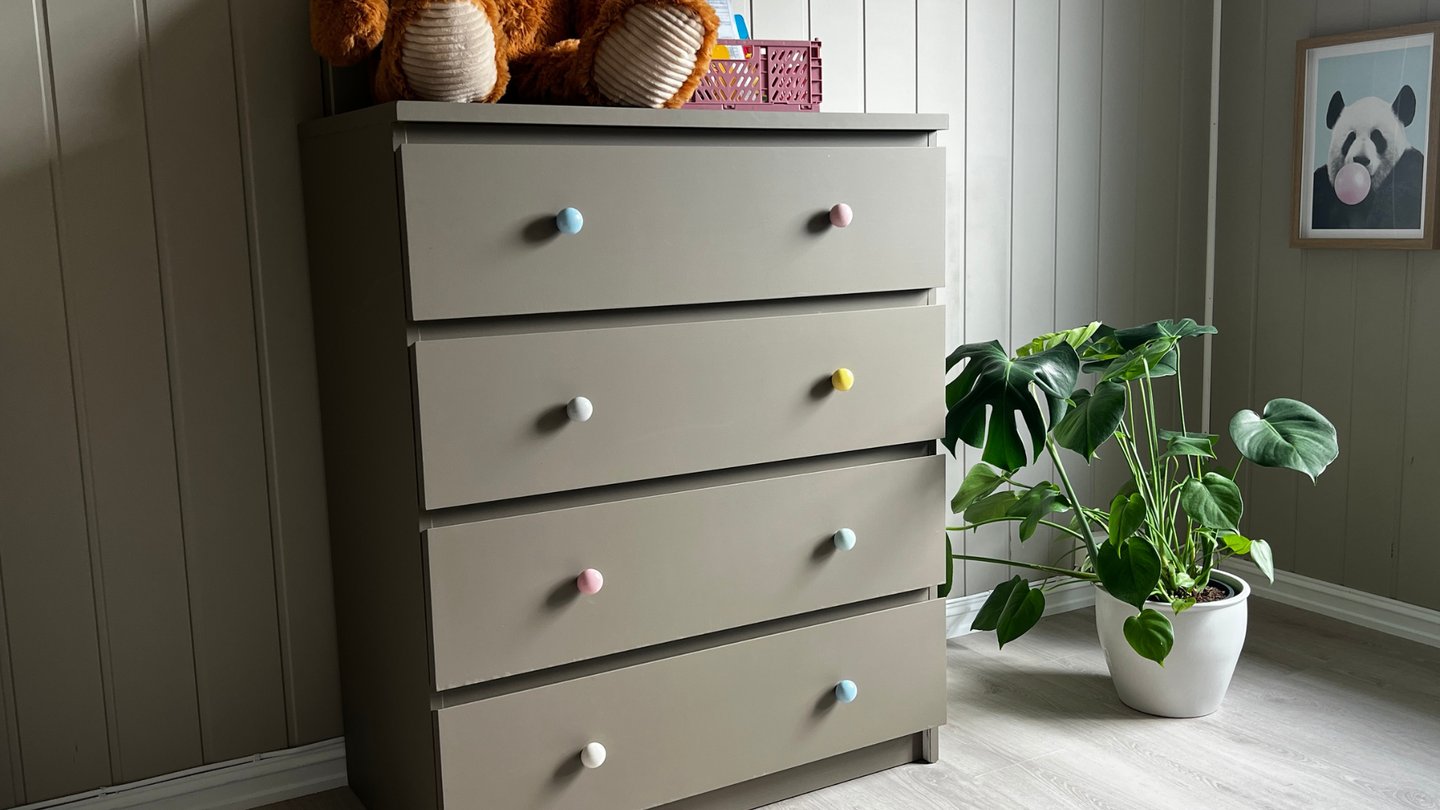 Ferdig kommode i brungrå farge med fargerike knotter og bamse på toppen.