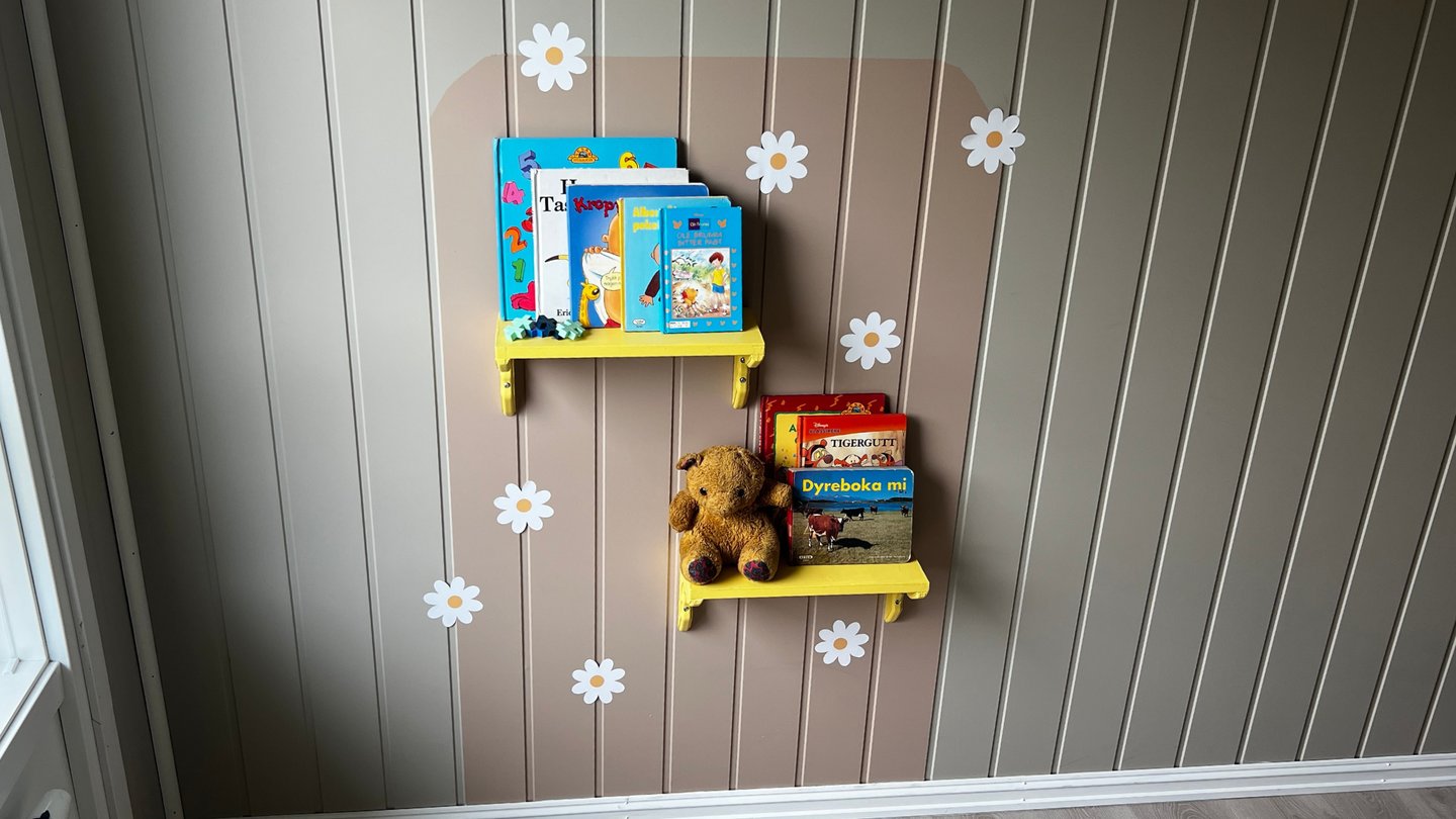 Ferdig lesekrok med gule hyller, bøker og bamse på vegg dekorert med blomster.