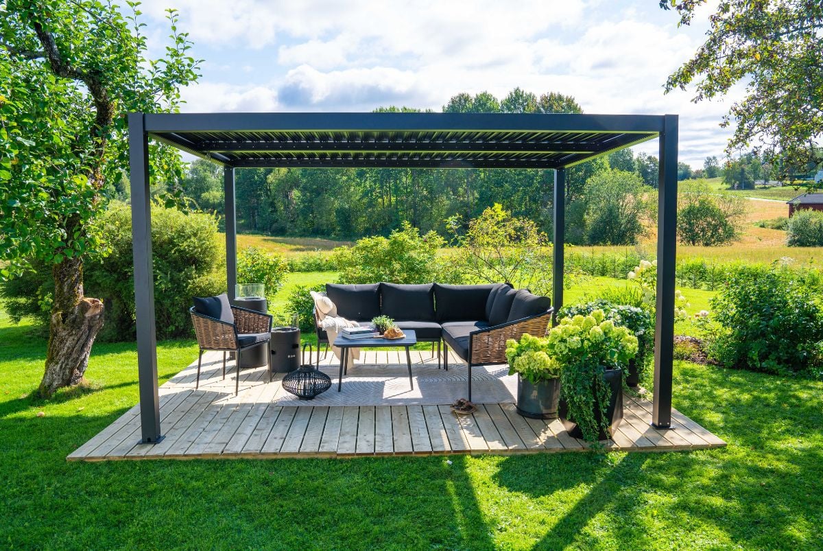 Bord og hjørnegruppe under sort pergola i stor hage med utsikt.