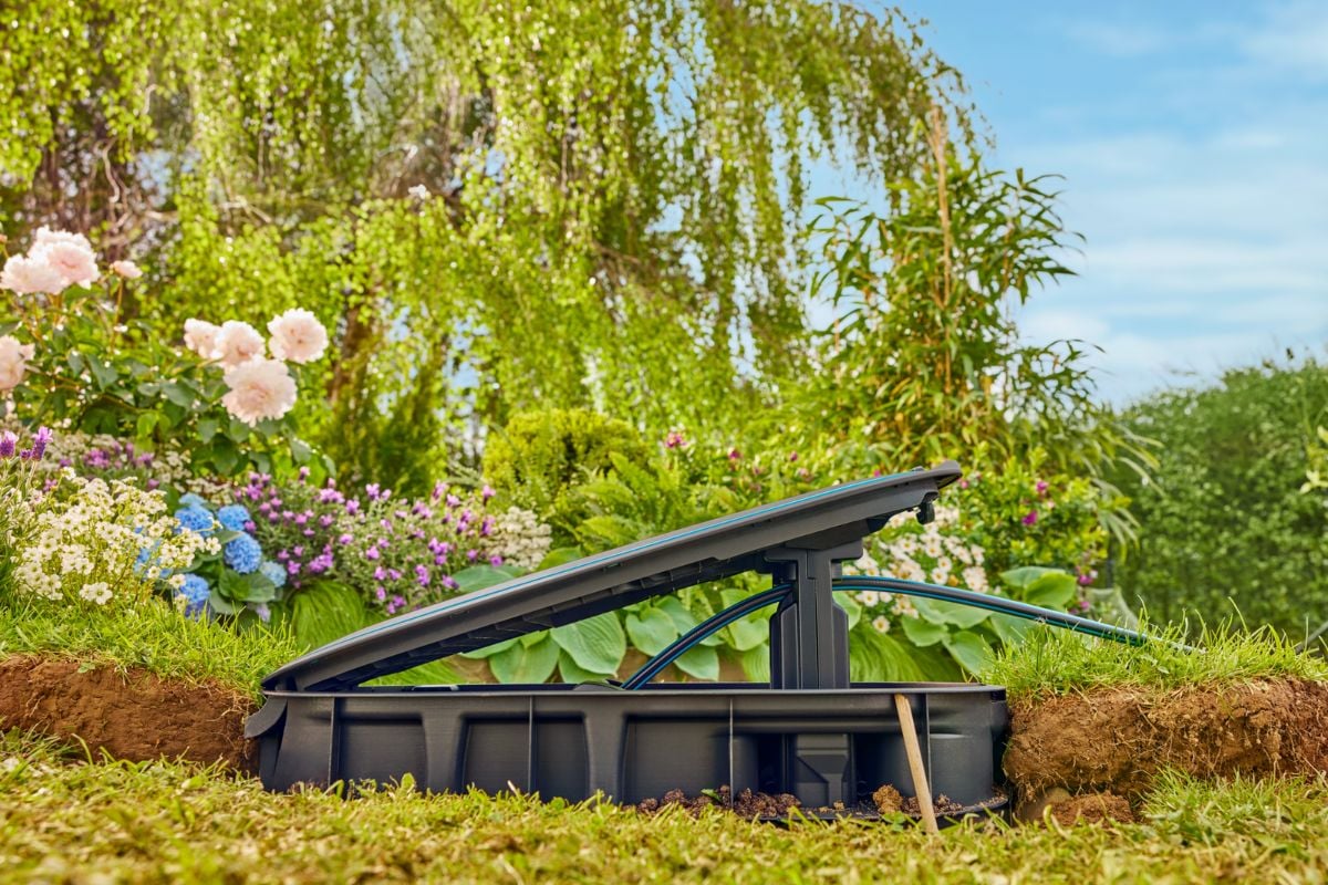 Åpen Gardena slangetrommel i plen, omkranset av blomster og trær