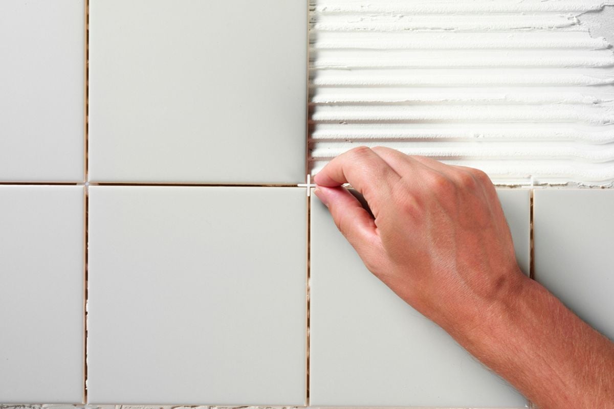 Hånd legger hvite keramiske fliser over lim på vegg.