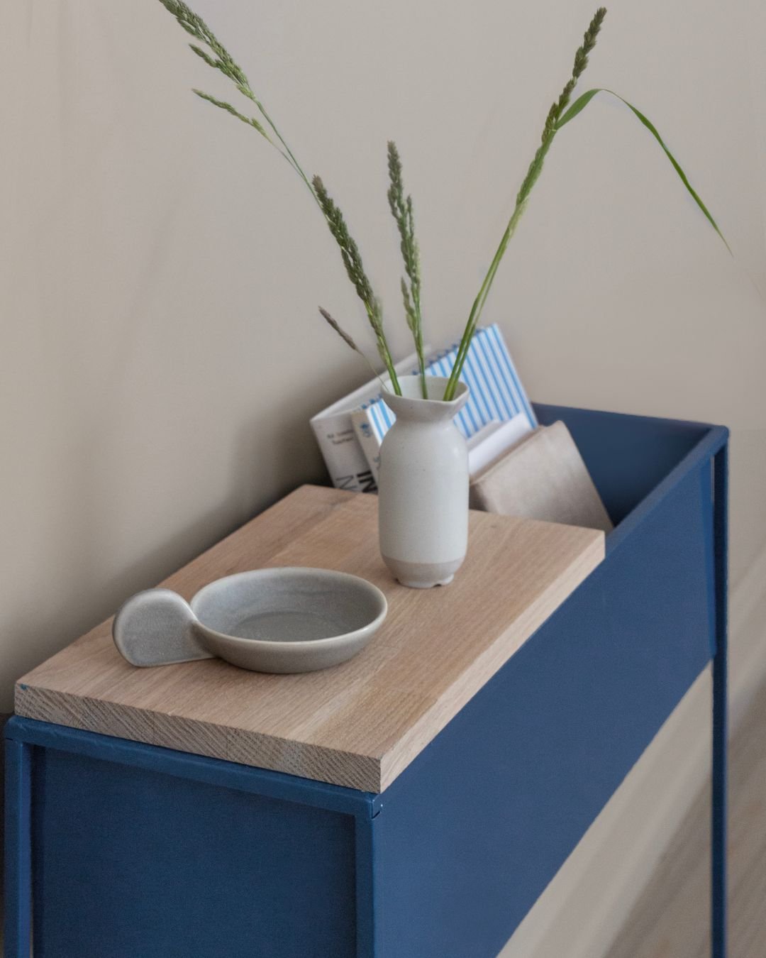 Blå skjenk med lys treplate, liten hvit vase med strå, skål og bøker stående bak.
