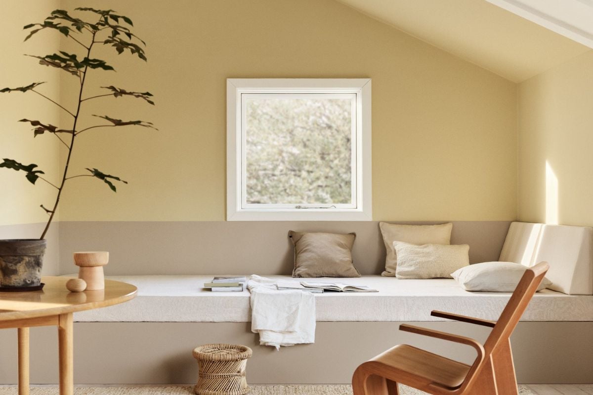 Lyst rom med gul og beige vegger, innebygd benk med puter, trestol og lite bord med plante.