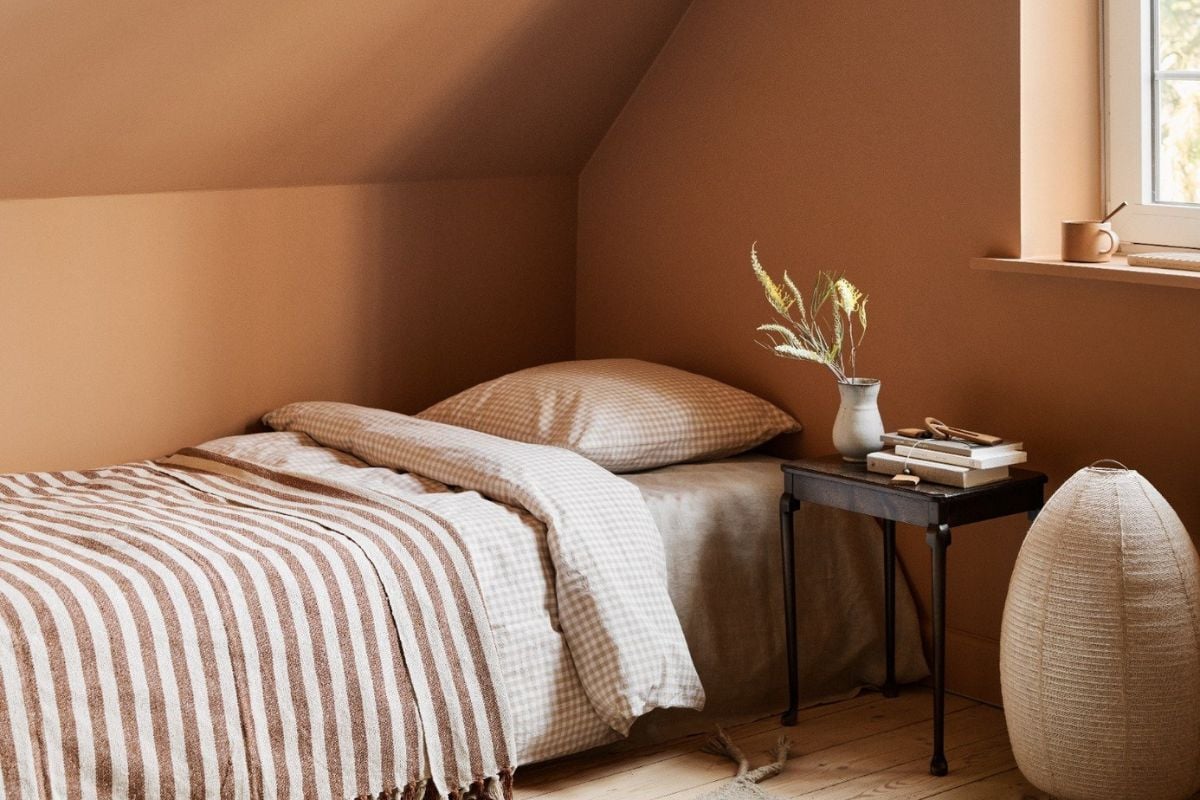 Lite soverom med skråtak og varme terrakottafargede vegger, enkel seng med stripet teppe og nattbord ved vinduet.