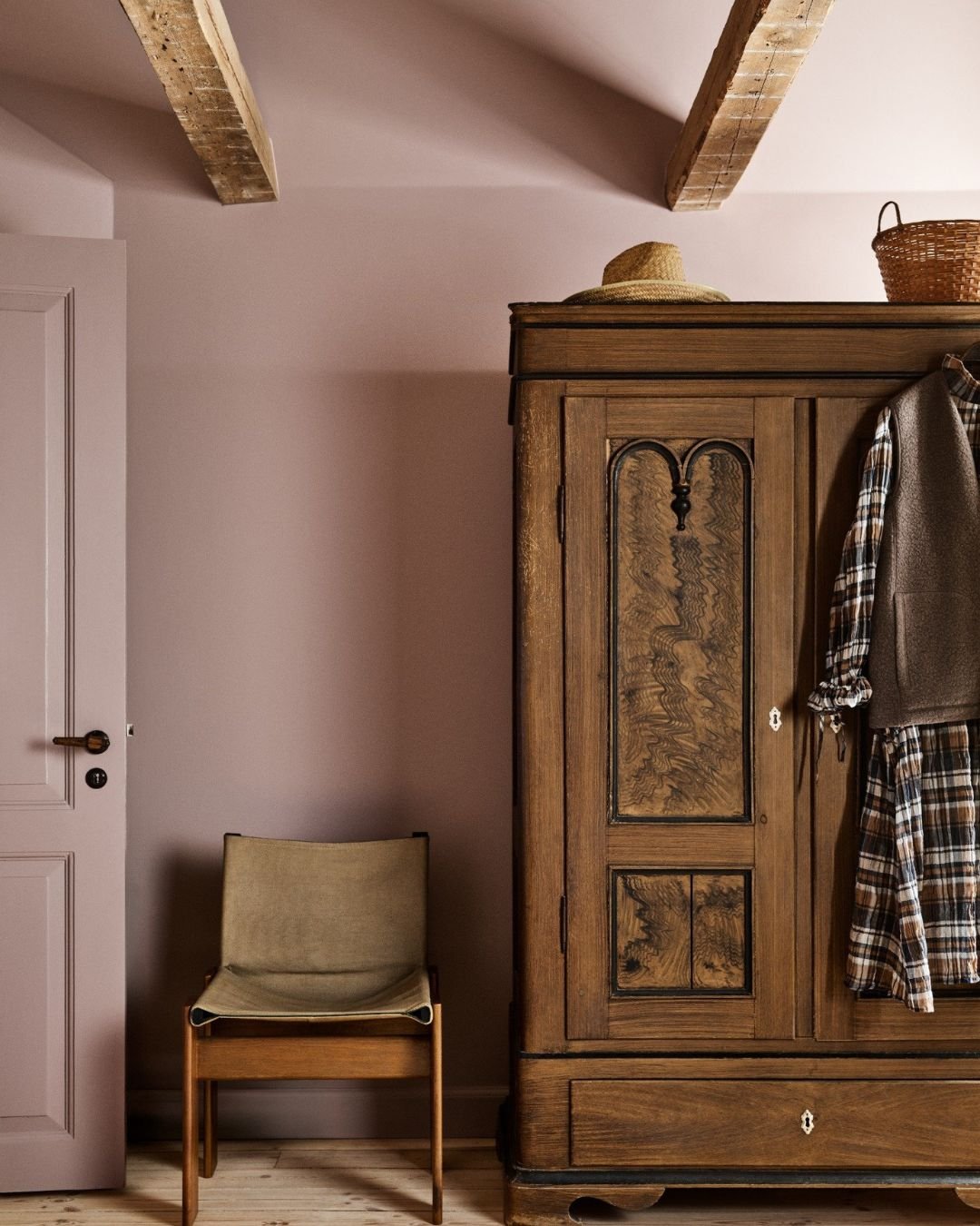 Rom med lys rosa vegger, gammel tre­garderobe, enkel stol og synlige bjelker i taket.