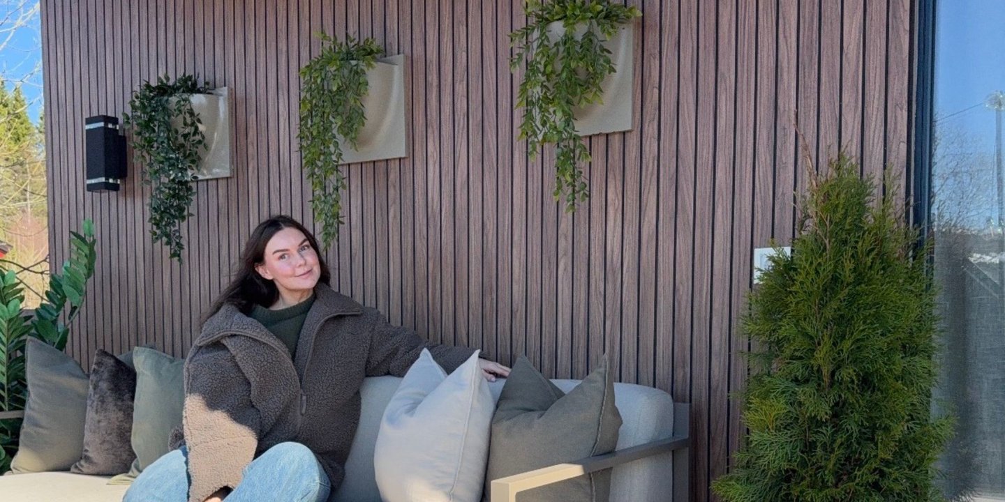 Kvinne sitter i utesofaen, med en spilevegg med blomster bak seg