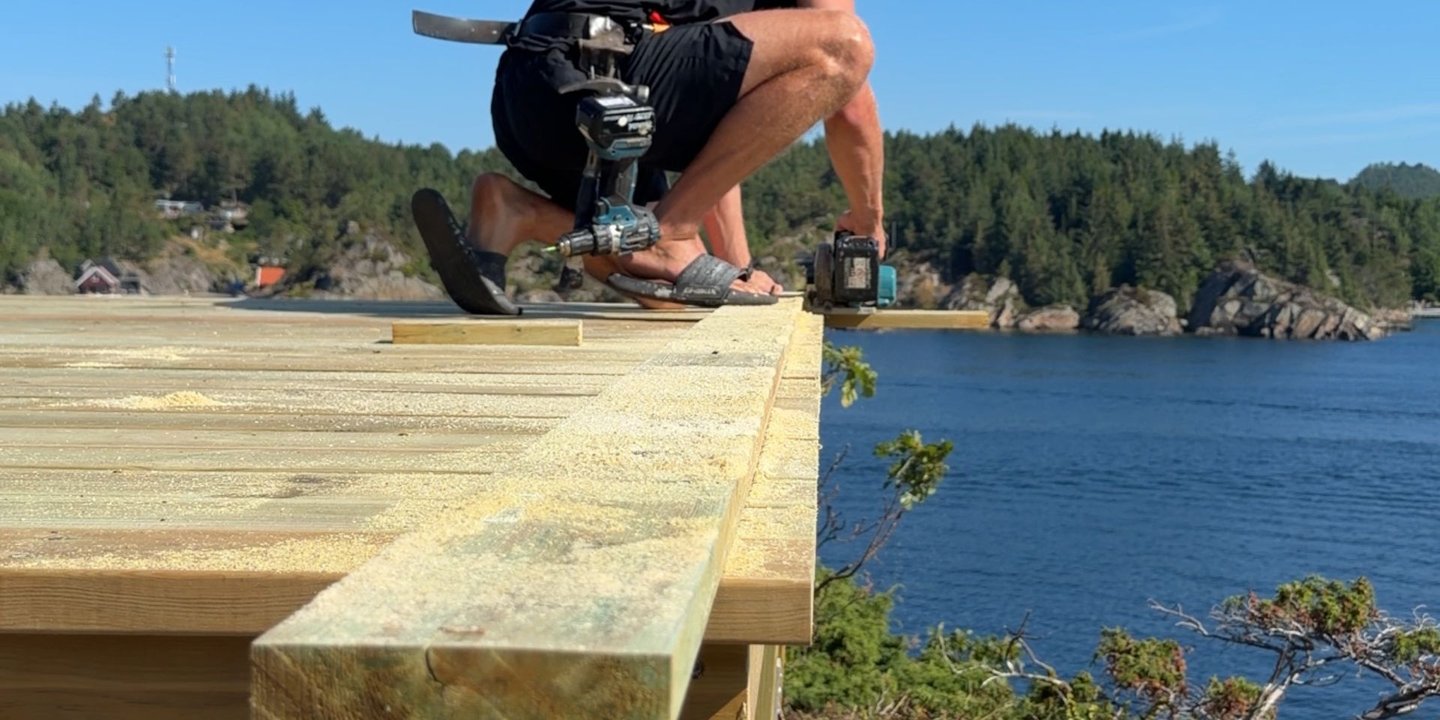 Gutt som kapper av overflødig treverk på terrassebordene i sommerlige omgivelser med havet i bakgrunnen