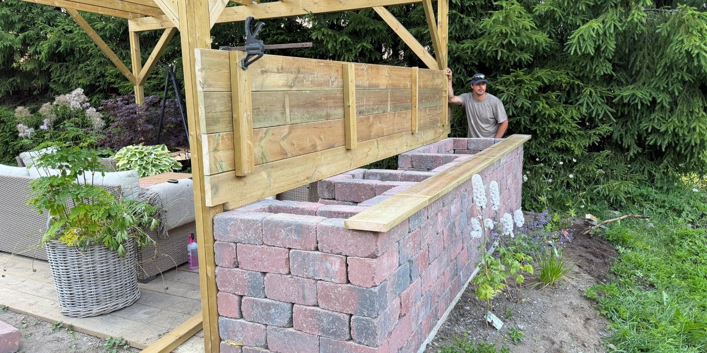 Montering av lokk i impregnert tre på utekjøkken bygget med murstein under pergola.