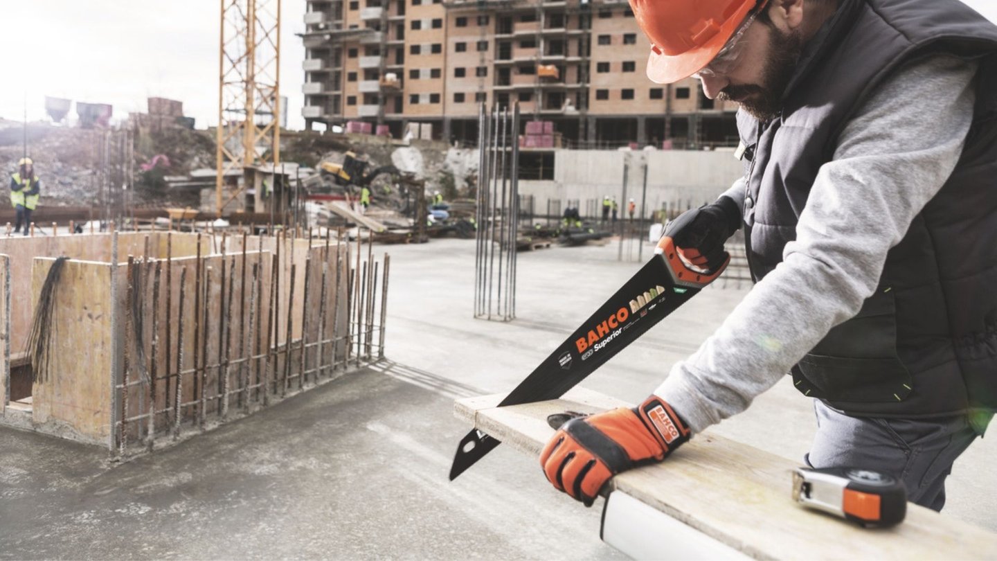 Mann med oransje vernehjelm står på byggeplass og sager med håndsag fra Bahco.
