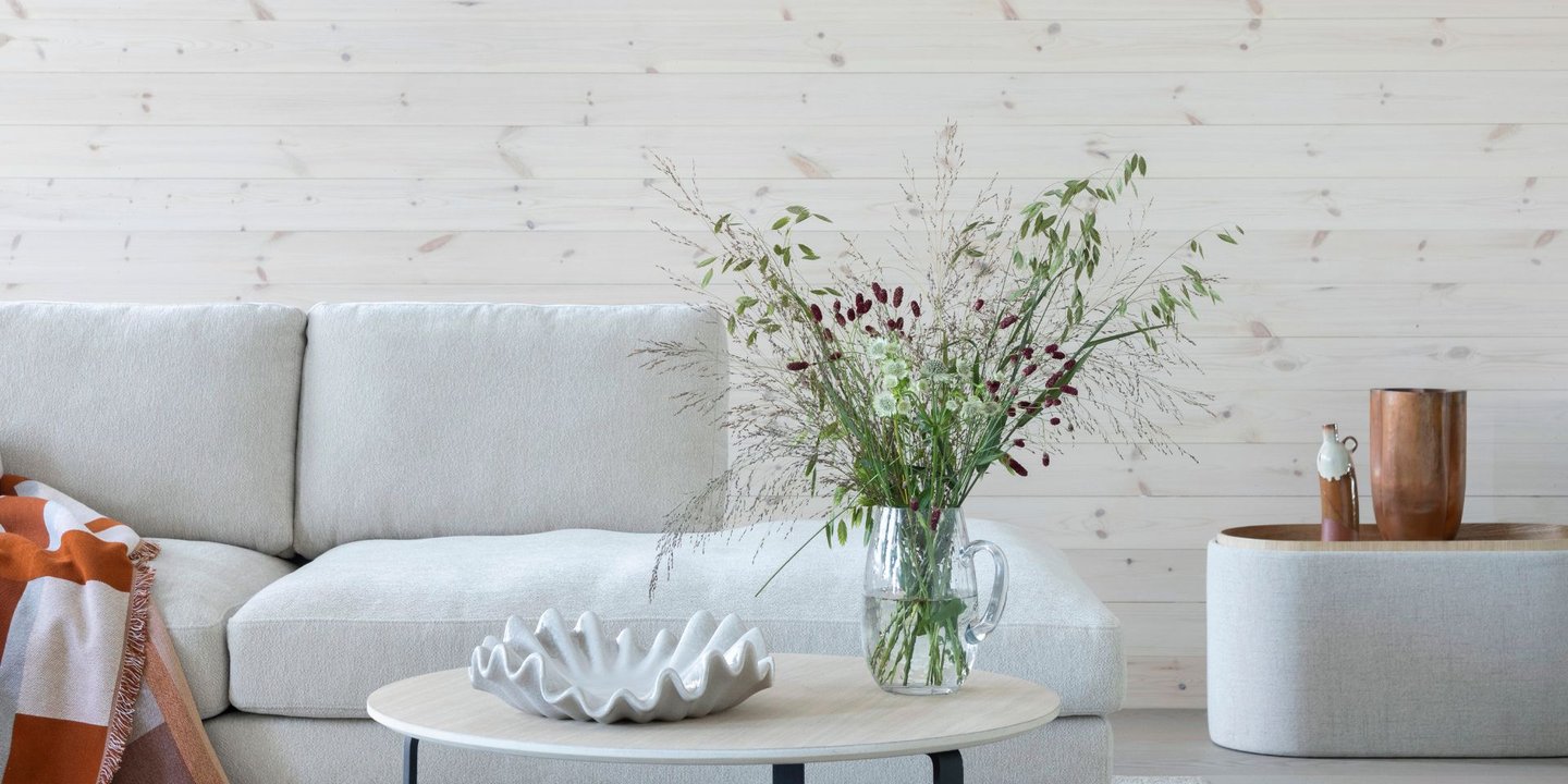 Lys stue med hvitbeiset trevegg, lys sofa og rundt sofabord. På bordet står en vase med blomster og en dekorativ skål.