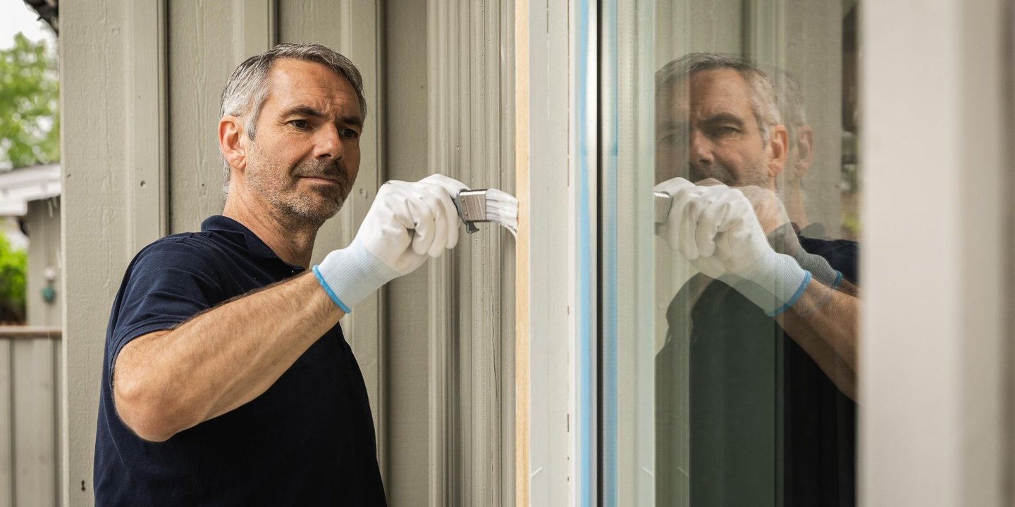 Mann maler vinduskarm på utsiden av et grått hus.