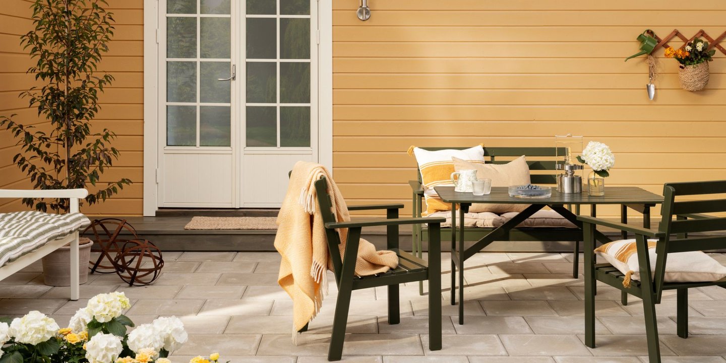 Et hus malt i gult med en mørk sittegruppe foran seg. Hvit glassdør med ruter inn til huset. Blomst i venstre hjørne.