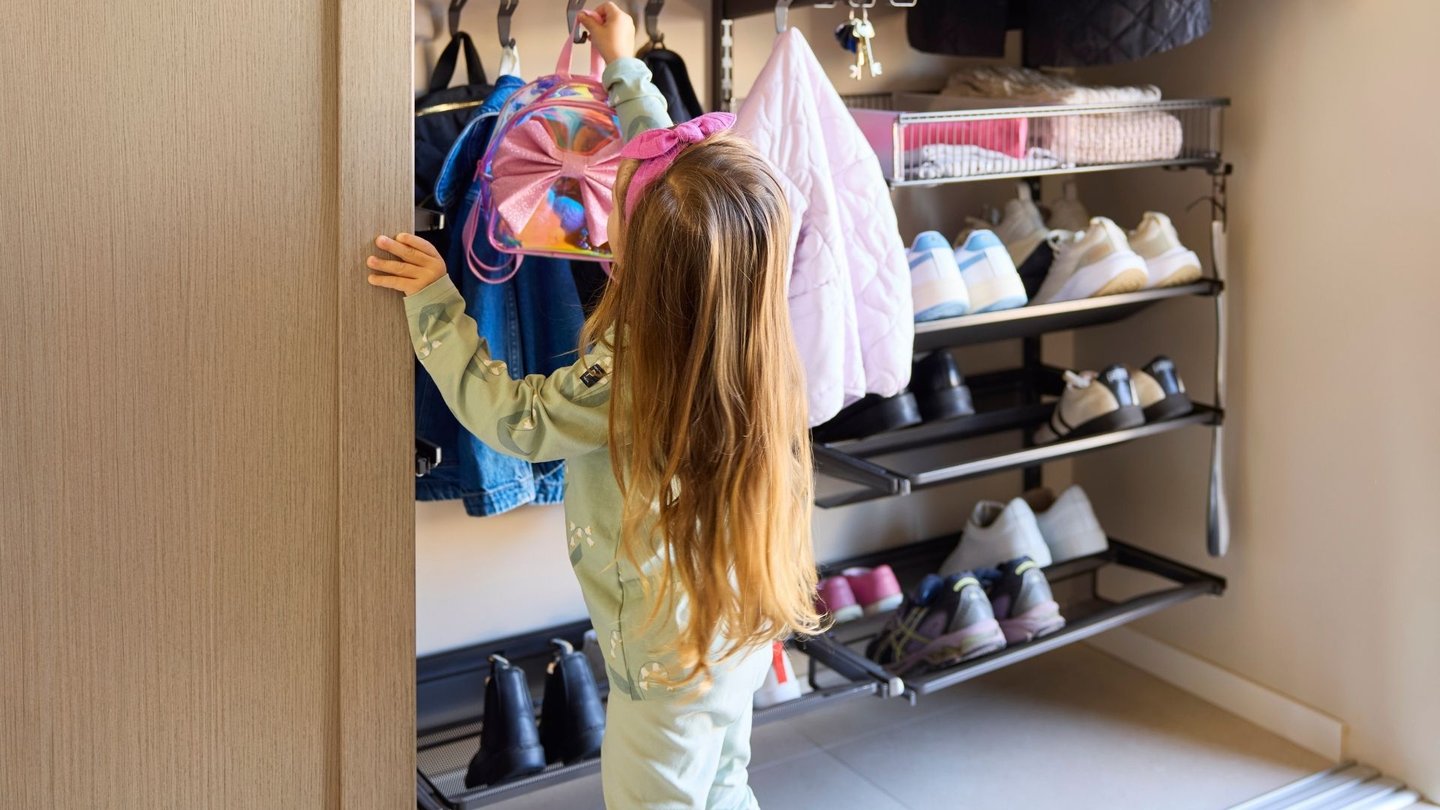 Liten jente i garderobe, med lav oppheng og skohyller, som strekker seg etter rosa sekk.
