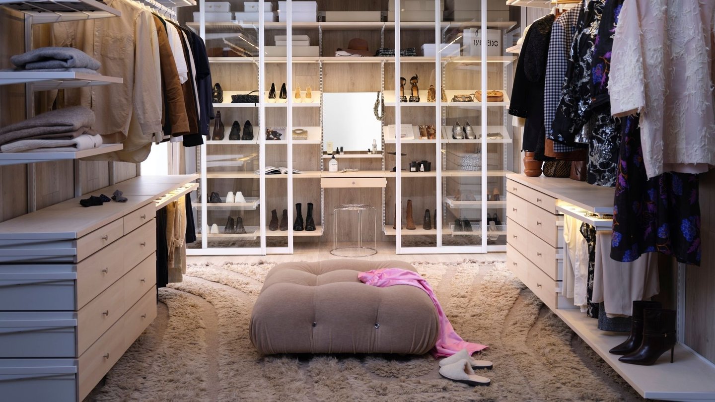 Bildet viser et moderne og ryddig walk-in-closet. Det har hvite skap og hyller med glassdører, fylt med klær, sko og tilbehør.