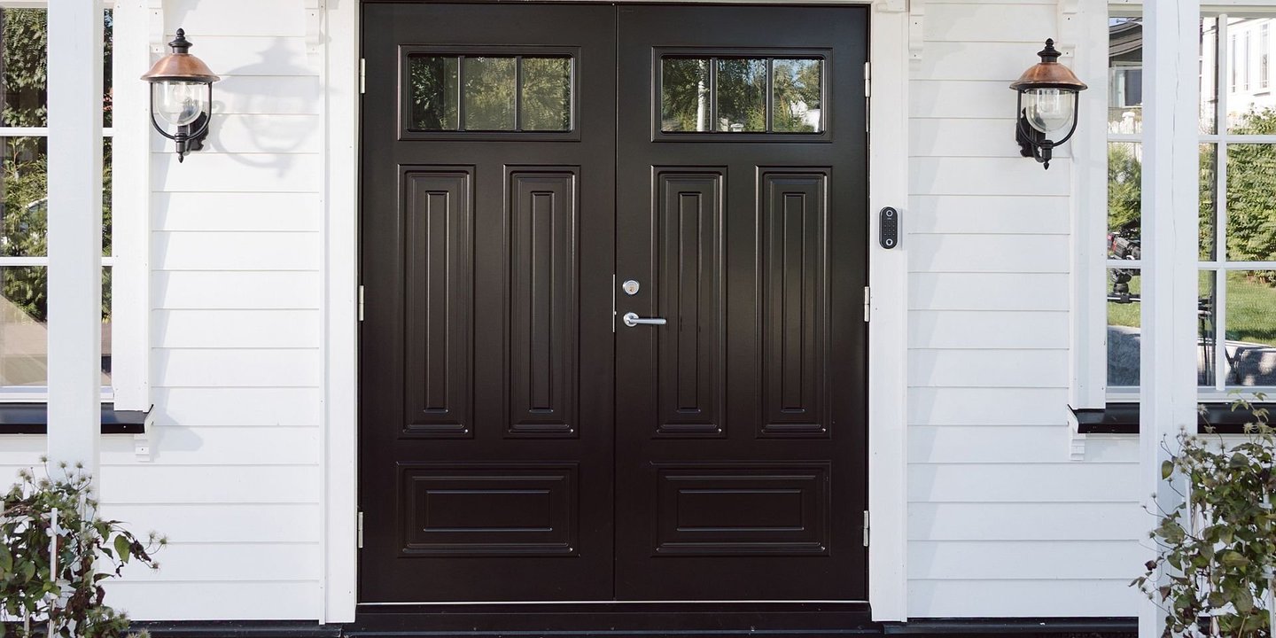 Svart doble inngangsdører med glassfelt øverst, montert på et hvitt trehus med to utendørslamper på hver side.»