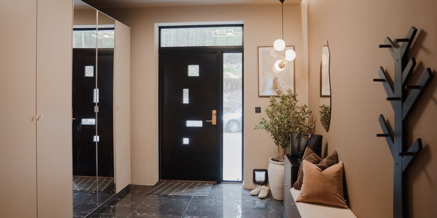 Bilde av en varm beige gang med en sort dør med vinduer i ulik størrelse. Det er pyntet med en benk med puter på og gulvet er flisbelagt.