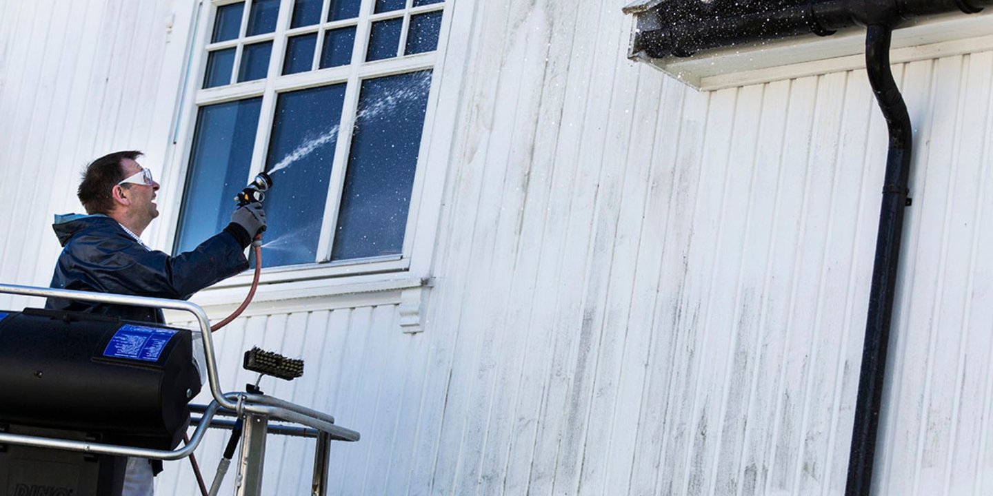 En mann står på en lift og vasker et skittent, hvitt hus med en høytrykksspyler.