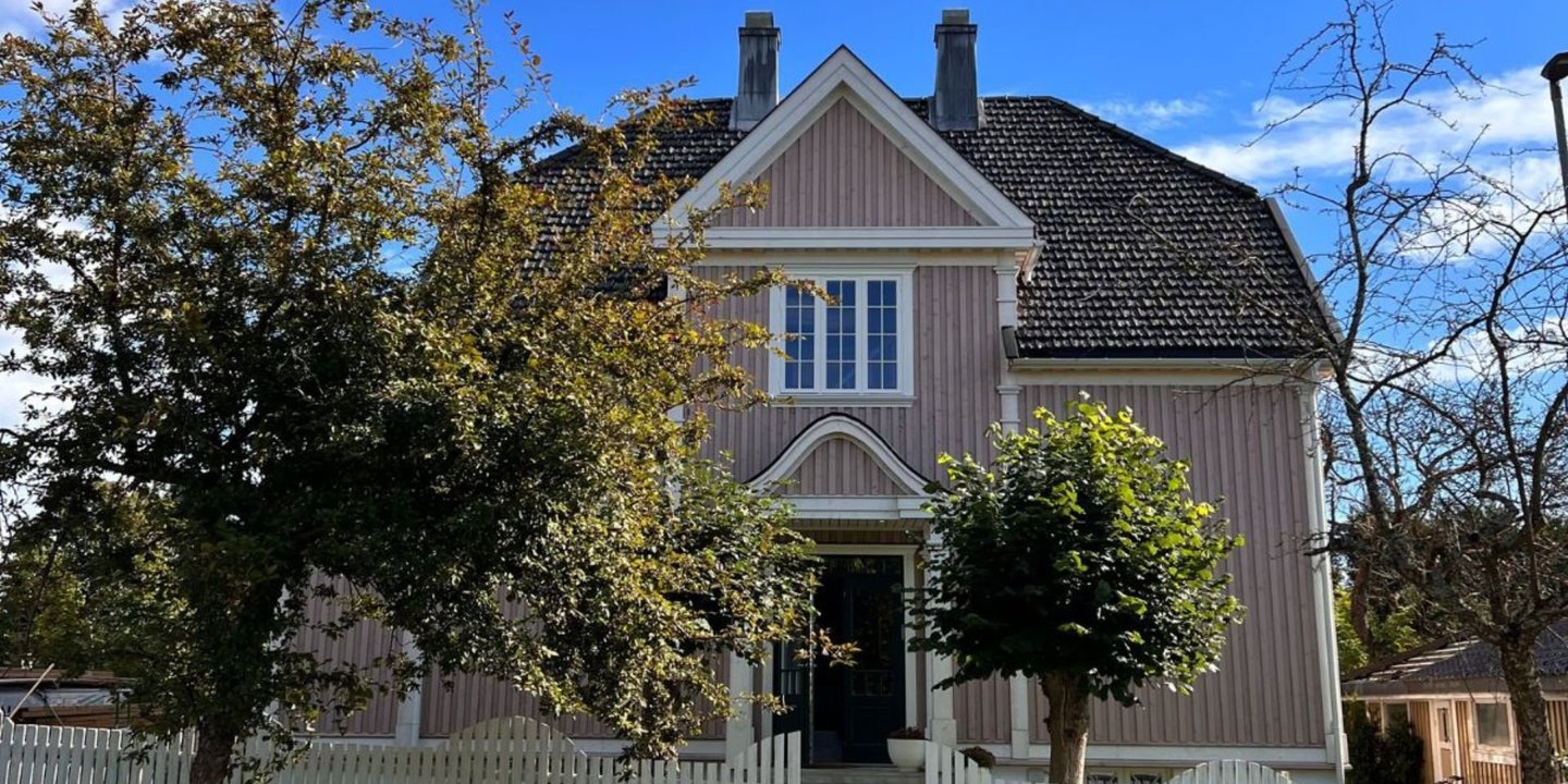 En gammel villa i dus rosa farge med hvite vinduer. Huset er omringet av grønne, frodige trær.