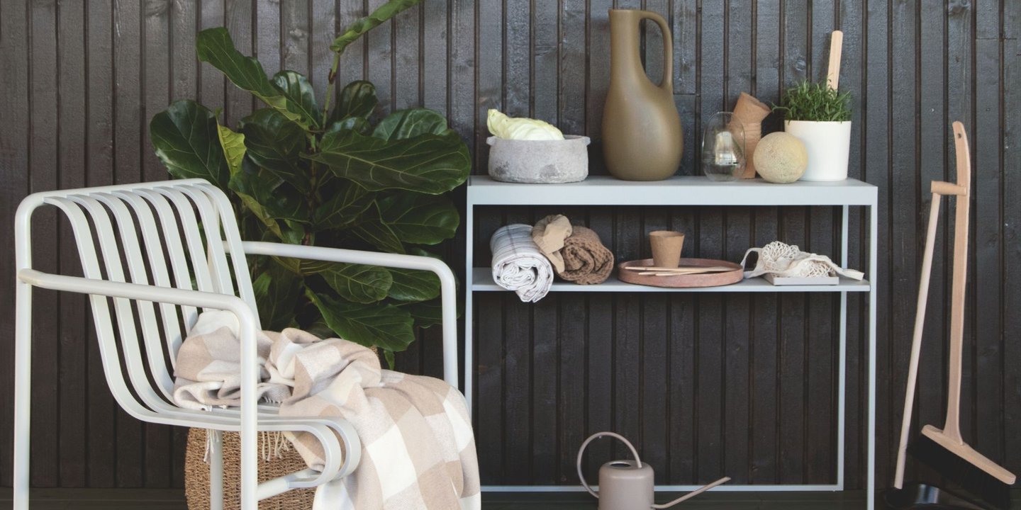 En lys stol med et pledd i seg foran en mørk beiset vegg. Det er også et konsollbord plassert ved siden av stolen, med en rekke pyntegjenstander. Det står også et kost- og støvbrett ved siden av konsollbordet og en grønn plante bak stolen.