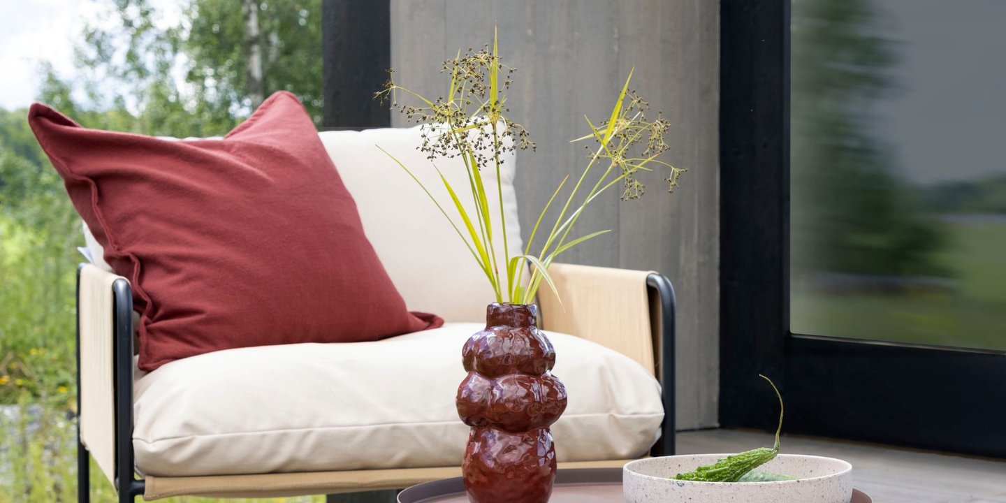 En loungestol med med en pute. Et lite avlastningsbord foran stolen med en vase med en grønn plante. Veggene på huset bak er i en rolig gråbeige farge.