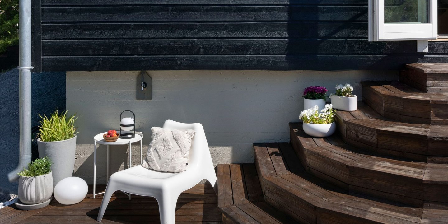 En brun trapp fra et svartbeiset hus ned på en brunbeiset terrasse. En hvit loungestol i høyre hjørne med planer og et avlastningsbord på siden av seg. En pute i stolen.