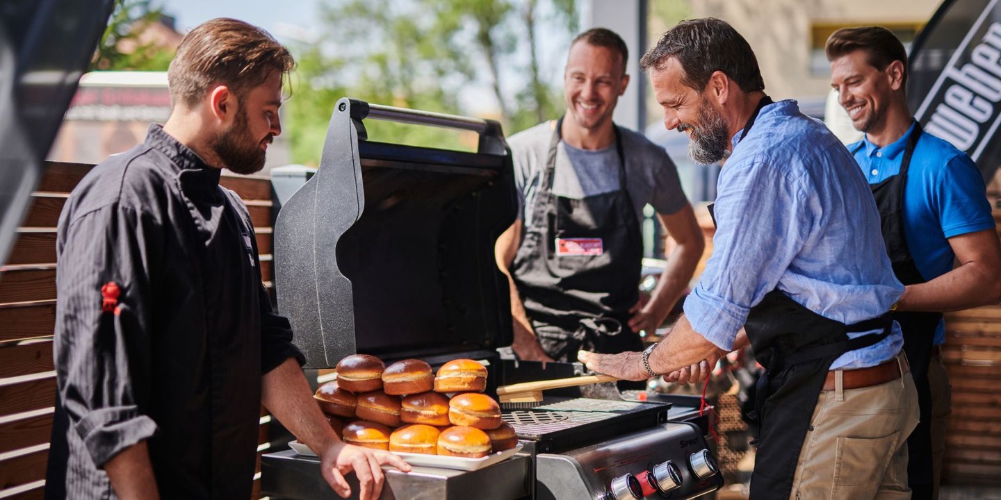 Fire menn står rundt en grill fra Weber mens de smiler. Hamburgerbrød står ved siden av grillen.