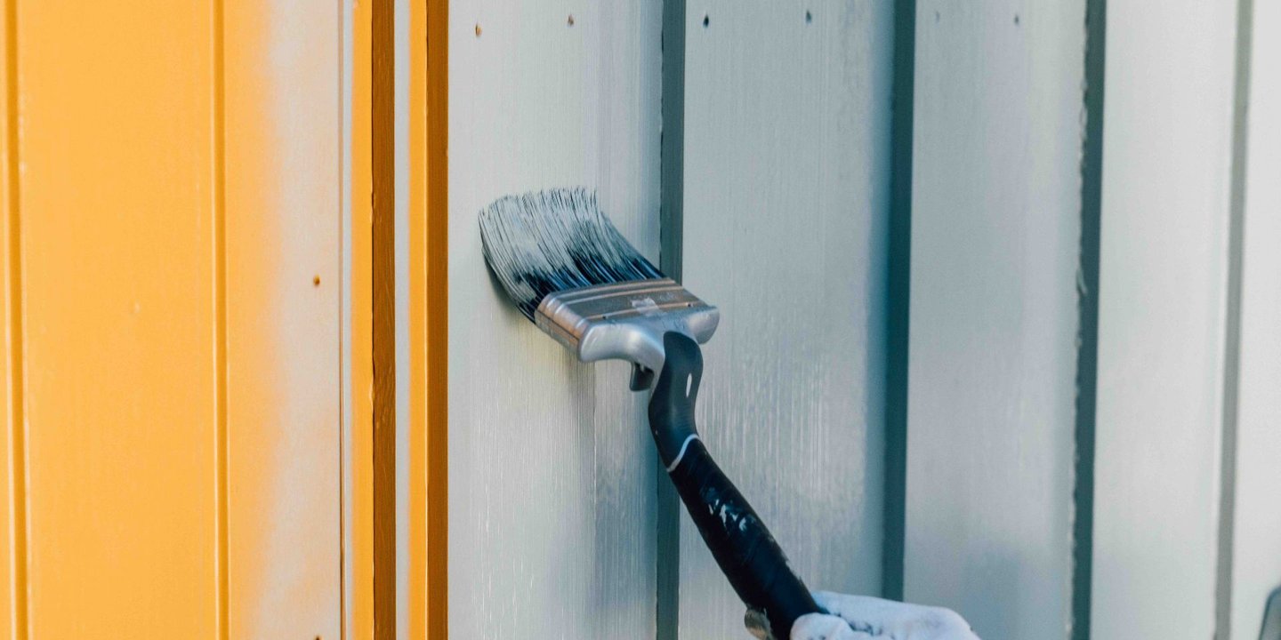 Person maler med grått på et gult hus med pensel