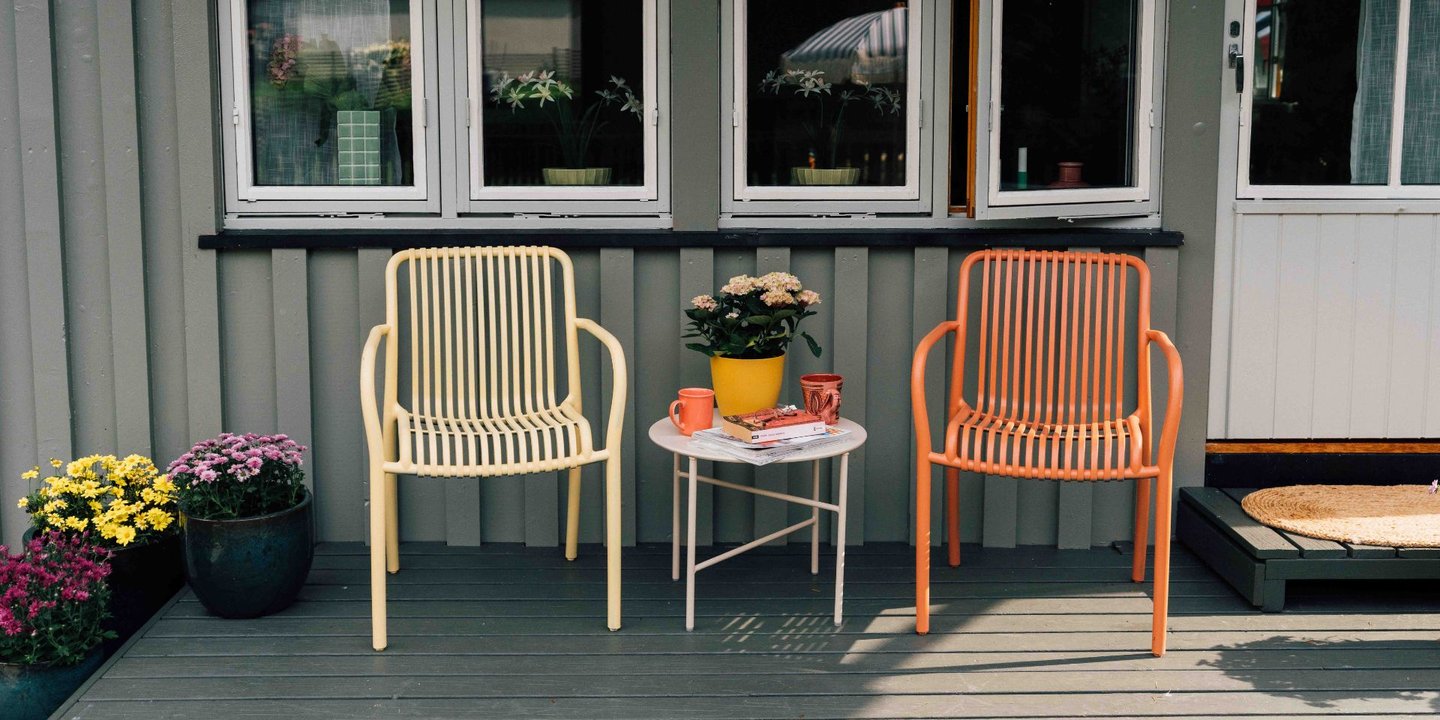 To stoler på en platting med blomster på venstre side. Det står også et lite bord mellom de to stolene. Den høyre stolen er oransje og den venstre er gul.