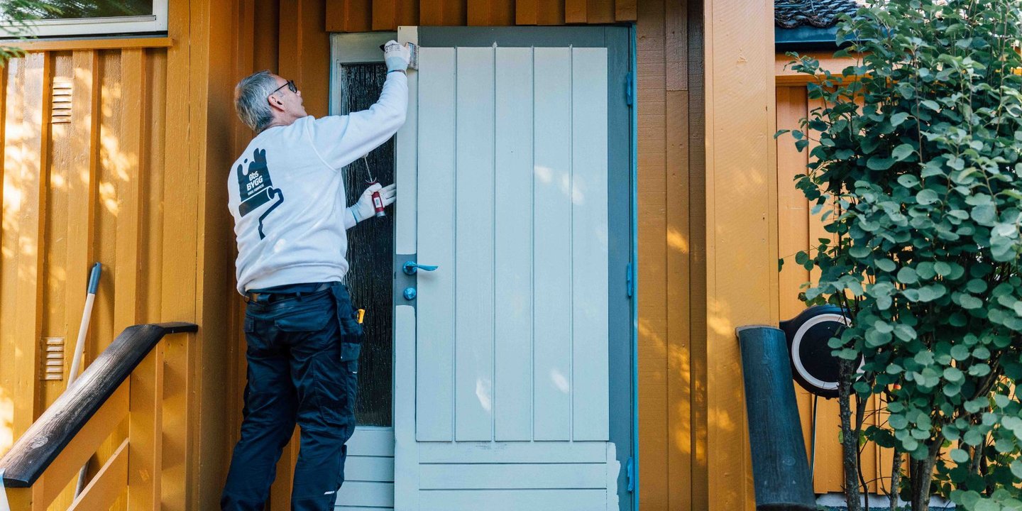 En mann i hvit genser maler en dør på et gult hus
