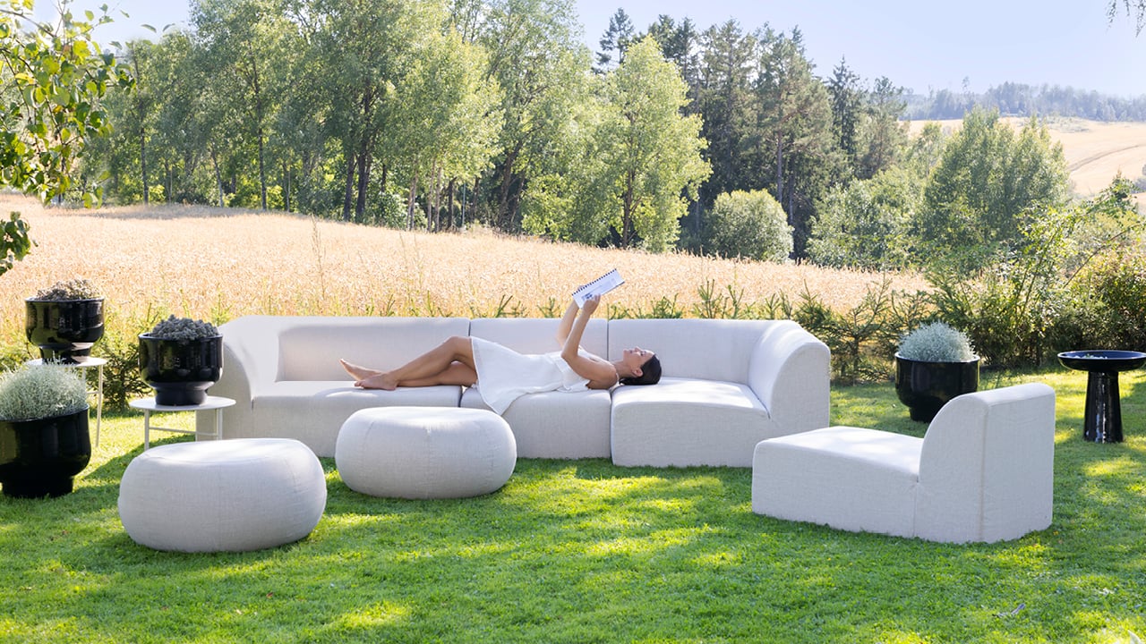 Skagen modulsofa i beige farge i en hage. En dame i hvit kjole ligger på sofaen og leser et magasin.