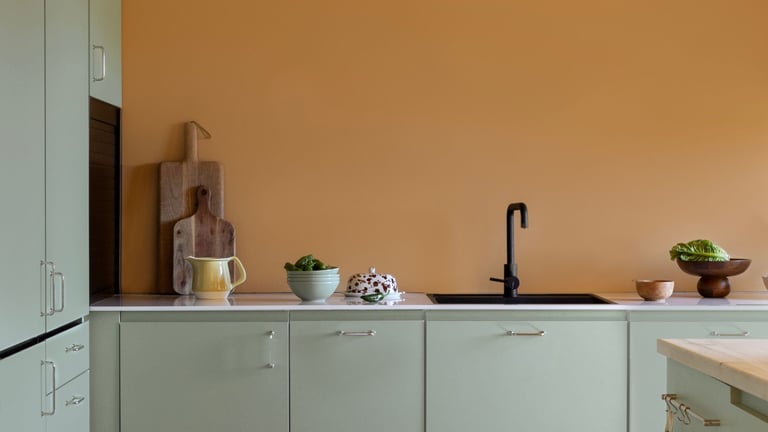 Kjøkken med oransje vegg, lysegrønne skap, svart kran og enkel dekor med boller og skjærefjøler.