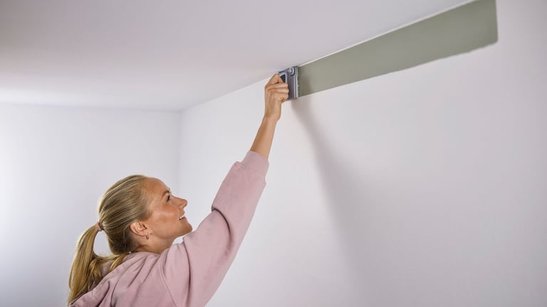 Kvinne maler øverst på veggen mot taket med en malepute, i fargen grå.