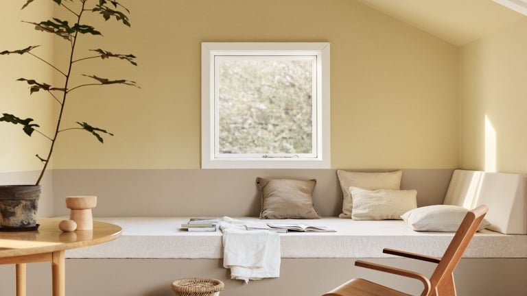Et hyggelig rom trestol, bord og en grønn plante. Veggene har en dempet farge av gul øverst og beige nederst. Det er innebygd sitteplass i hjørnet av rommet.