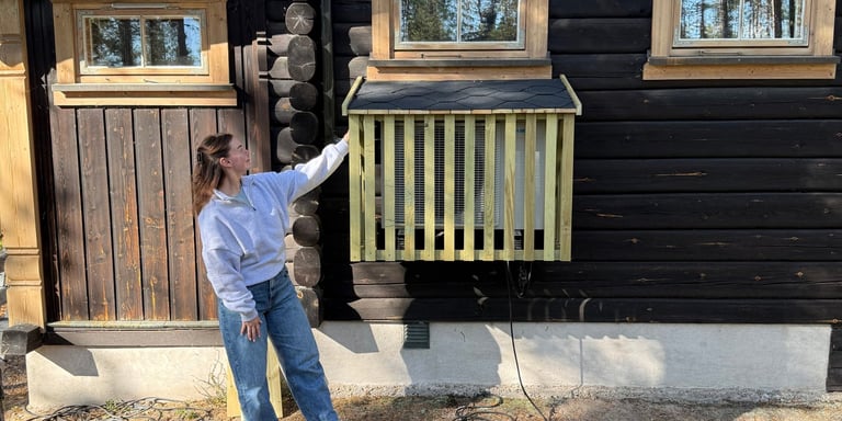Kvinne står foran hytta og peker på selvbygget varmepumpehus