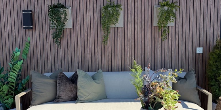 Uteområde på terrasse med vegg av spileplater med blomster. Foran står en en utesofa og utebord