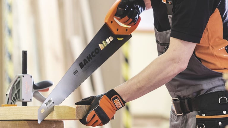 Mann i arbeidsklær og vernehansker som sager en treplanke med en Bahco håndsag på et arbeidsbord i et verksted.