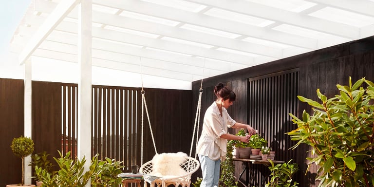 En kvinne står på uteplassen under trapestak fra gop, og tar på urter i potter.